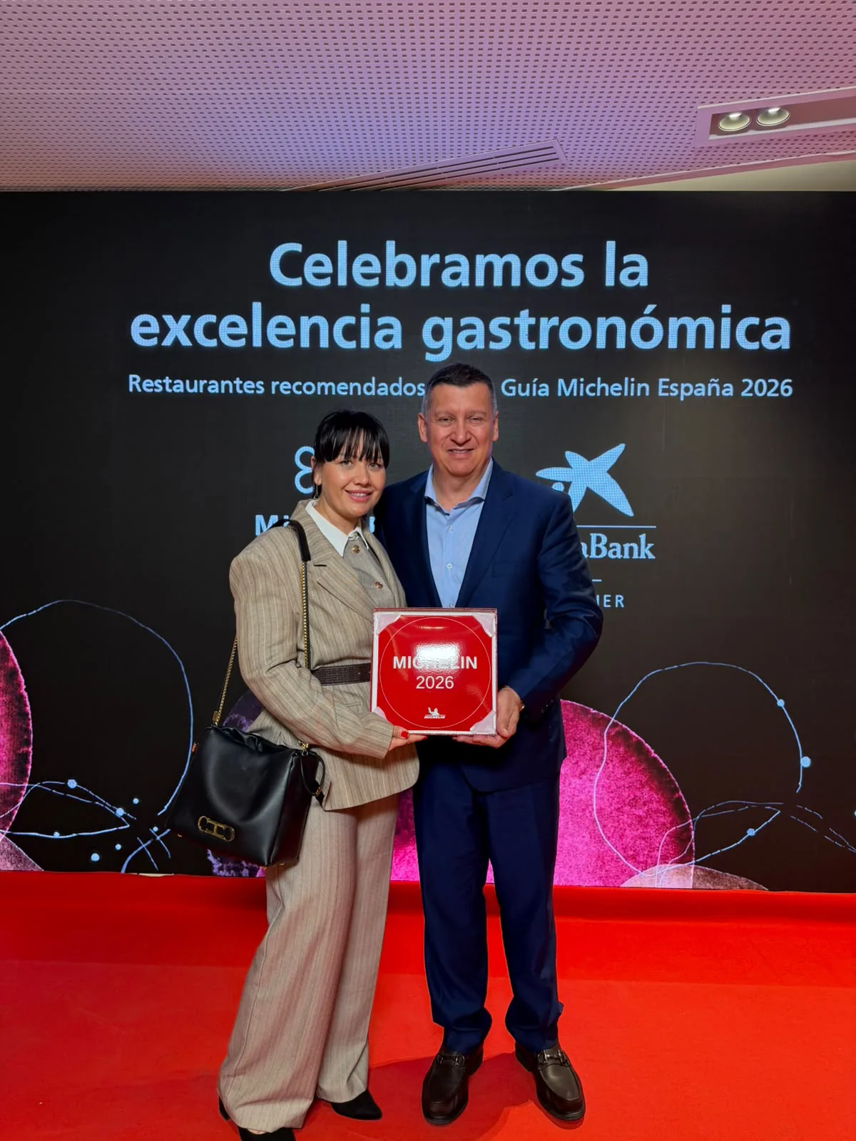 Pedro Nel im blauen Anzug und eine Frau im hellen Anzug halten die rote Plakette MICHELIN 2026 vor der Tafel der empfohlenen Restaurants des Guide Michelin Spanien 2026