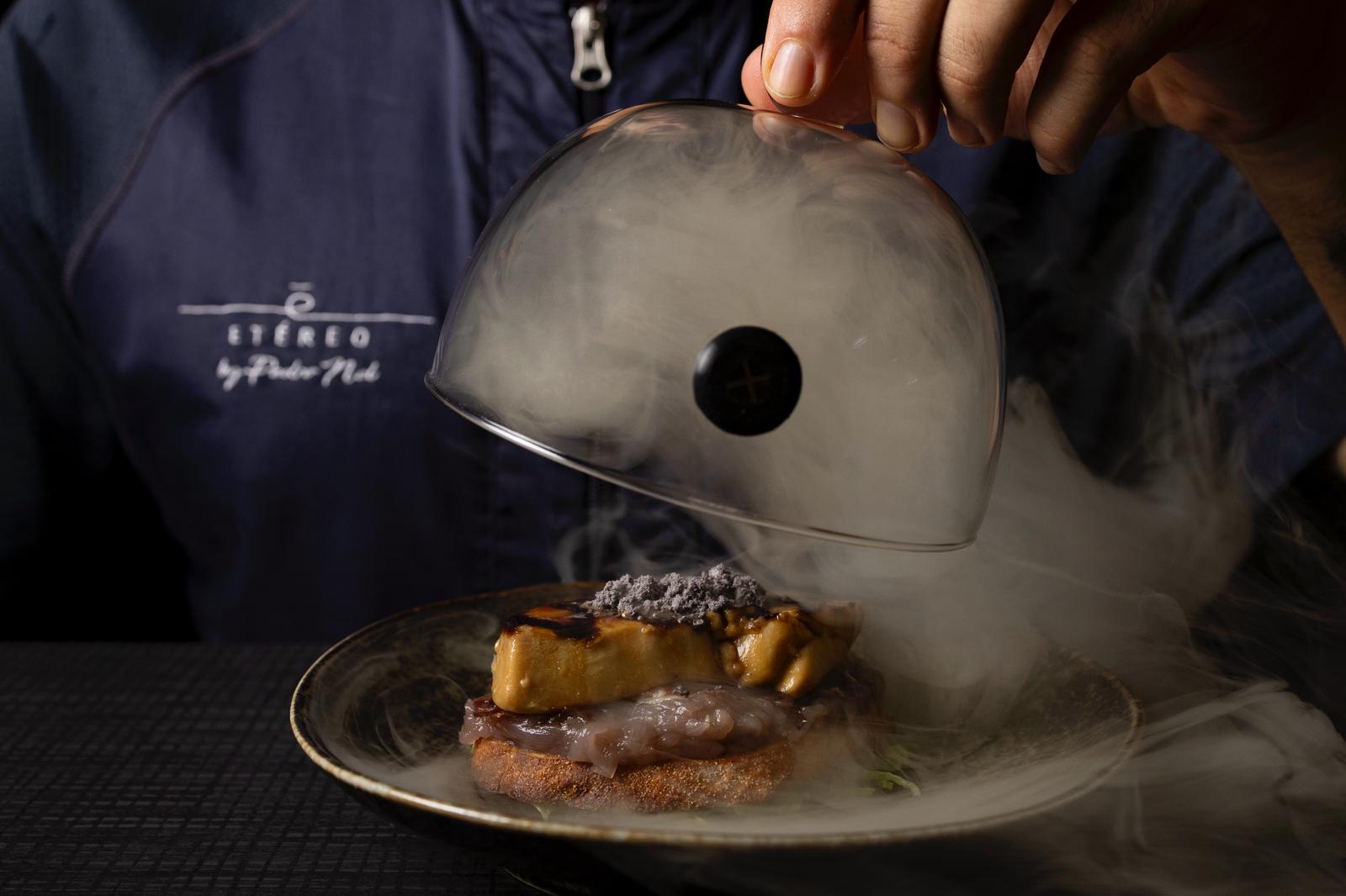 Una mano levanta una campana de cristal y deja salir el humo sobre un plato