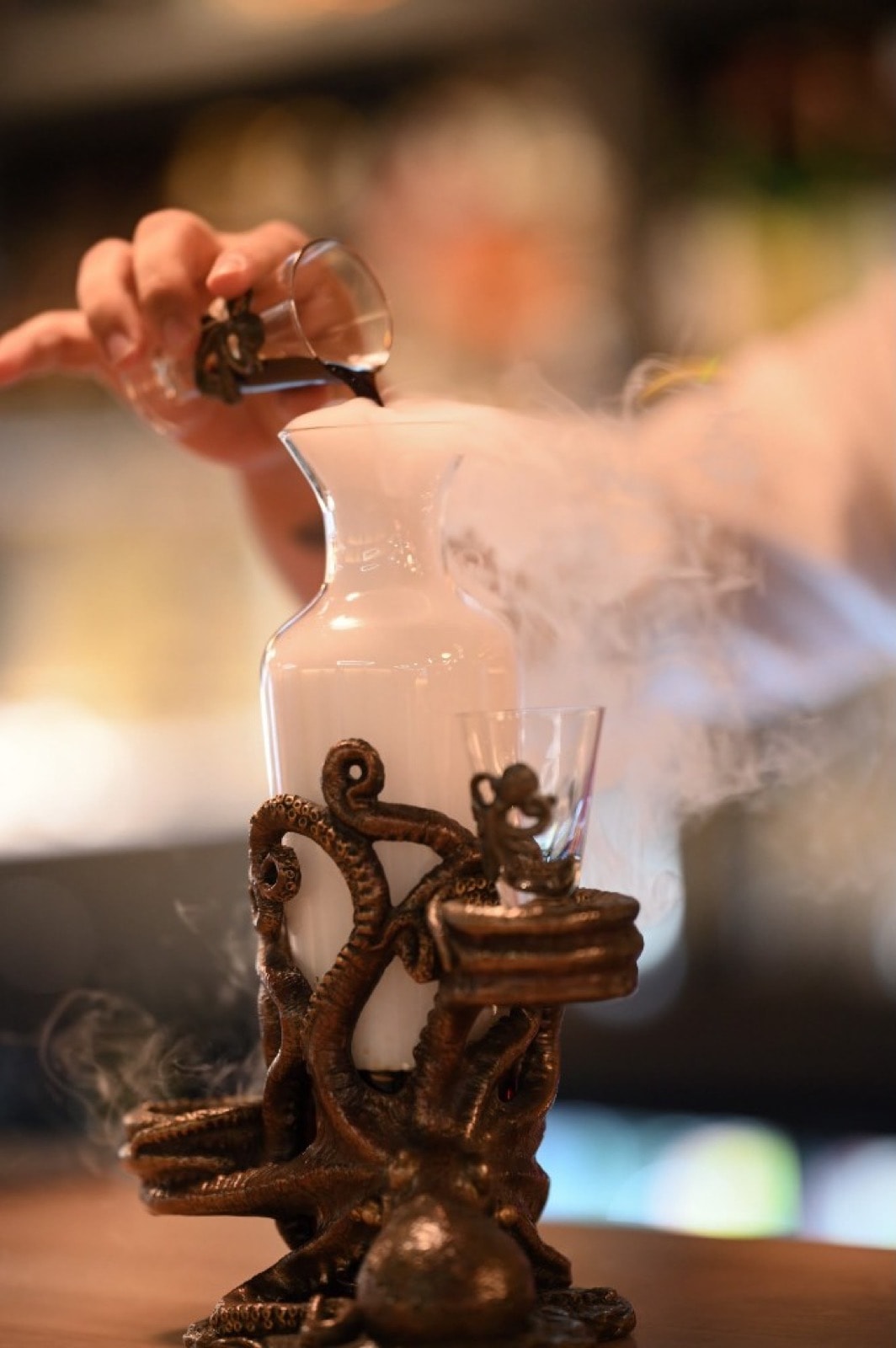 A bartender pours a dark liqueur into a glass jug overflowing with dry-ice vapour, on a bronze octopus-shaped stand