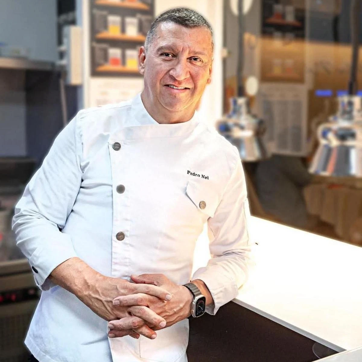 Pedro Nel sonríe en su cocina, con la chaquetilla blanca bordada con su nombre y las manos entrelazadas, apoyado en la encimera